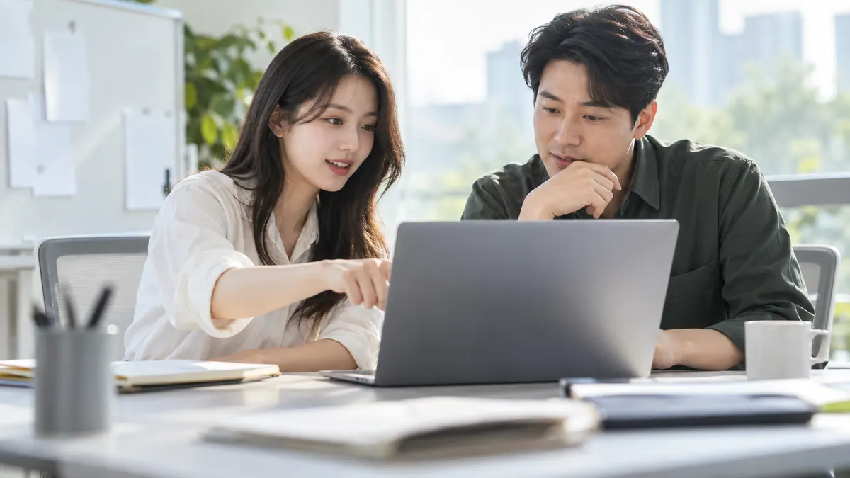 마케팅 담당자 두 명이 회의실에서 노트북 한 대를 함께 보며 검색 결과에 나갈 문구를 고르는 기획 이미지